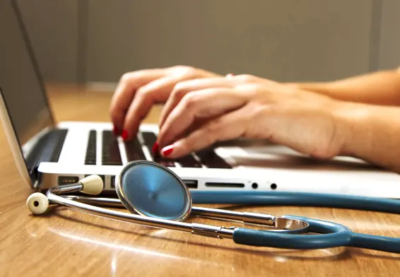 A healthcare professional typing on a laptop with a stethoscope placed on the desk
